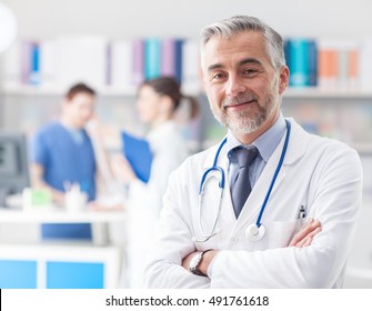 Confident smiling doctor posing and looking at camera with arms crossed, medical staff working on the background