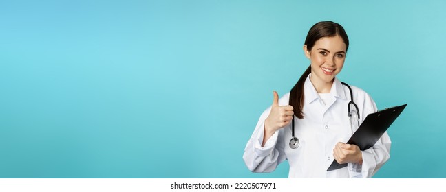 Confident Smiling Doctor Woman, Physician Showing Thumbs Up, Holding Clipboard, Appointment In Hospital, Standing Over Torquoise Background.