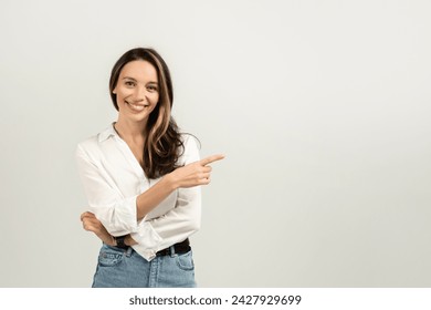 Confident and smiling european young brunette woman in a white blouse pointing to the side while standing against a plain light background with arms crossed, studio. Work, business ad and offer - Powered by Shutterstock