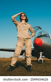 Confident smiling female pilot poses in front of the plane looking into the sky. Commercial and military aviation. Full length portrait on the airfield in the background of a blue sky.