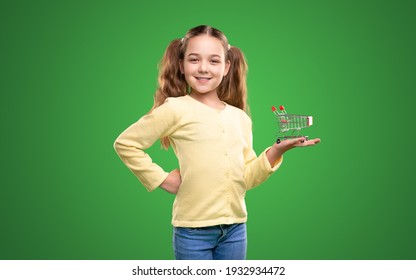 Confident smiling girl in casual clothes showing mini toy shopping cart on hand while representing supermarket against green background