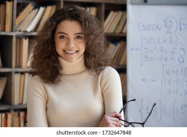 Confident smiling hispanic young woman math teacher, happy latin woman college student, professional tutor looking at camera standing arms crossed in classroom with whiteboard. Head shot portrait.