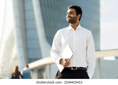 Confident smiling Indian businessman wearing white shirt holding laptop looking away on urban street. Successful business - Powered by Shutterstock