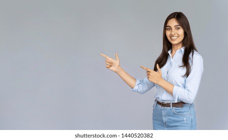 Confident smiling indian young woman student wear blue jeans shirts looking at camera pointing at copy space isolated on grey studio background, happy lady professional showing aside portrait, banner