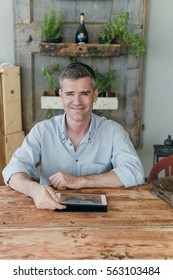 Confident smiling man using a digital tablet on a rustic wooden table, he is looking at camera