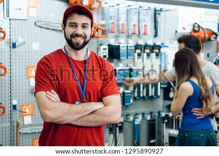 Confident smiling salesman on foreground in power tools store. Guy is ready to help clients on background.