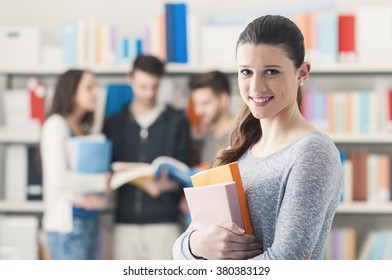 Confident Smiling Student Girl Posing In The Library, Holding Books And Looking At Camera, Learning And Education Concept