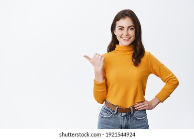 Confident smiling woman in yellow sweater, pointing finger left and showing advertisement on white copy space, standing against white background. - Shutterstock ID 1921544924
