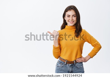 Confident smiling woman in yellow sweater, pointing finger left and showing advertisement on white copy space, standing against white background.