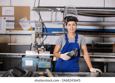 Confident smiling young woman in blue overalls, protective face shield and gloves standing with handheld angle grinder at profile cutting table in window production and assembly workshop.. - Powered by Shutterstock