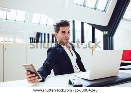 Confident smirking young man in formal suit relaxing from work checking social media using mobile in modern office looking at camera