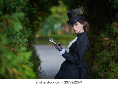 A confident and sophisticated lady in a strict black suit of the 19th century poses with a walking stick against the backdrop of an autumn park.