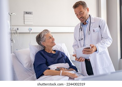 Confident specialist holding digital tablet and talking to old patient. Happy professional friendly doctor meeting elderly patient on a hospital bed and explaining health results. - Powered by Shutterstock