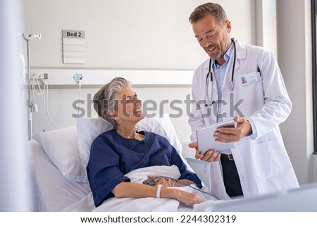 Confident specialist holding digital tablet and talking to old patient. Happy professional friendly doctor meeting elderly patient on a hospital bed and explaining health results.
