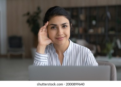Confident specialist. Portrait of motivated young indian woman expert worker scientist sit by laptop look at camera above screen. Smiling young woman pose for photo at desk distracted from work online