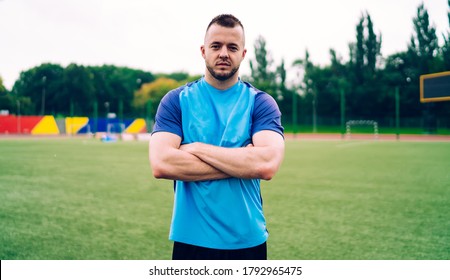 Confident Sportsman In Blue T Shirt Standing With Arms Folded Looking At Camera On Green Sports Field On Summer Day