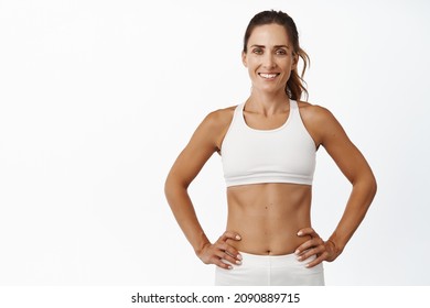 Confident Sportswoman In White Sportsbra, Holding Hands On Waist, Fitness Trainer Standing In Power Pose, Workout In Gym, White Background