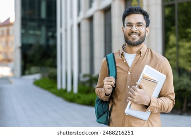 Confident student standing outside modern university building. Young man wearing glasses, carrying backpack, holding books and laptop. Concept of education, college life, academic success, study