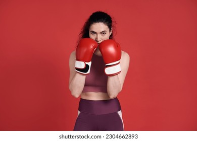 Confident studio portrait of young active determined strong female athlete, sportswoman boxer fighter posing isolated over red background in boxing gloves. People. Sport. Competition. Battle. Fighting - Powered by Shutterstock