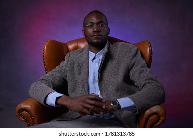 Confident stylish African American businessman sitting on a luxurious leather sofa on a dark background illuminated by red and blue light - Powered by Shutterstock