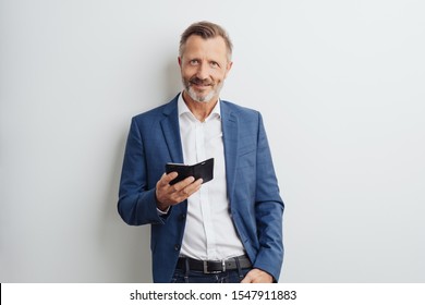 Confident Stylish Businessman Holding A Mobile Phone In His Hand As He Looks Up At The Camera With A Quiet Smile Against A White Studio Wall