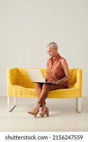 Confident Stylish Businesswoman With Tattoo Sitting On Sofa And Using Laptop, Studio Shot