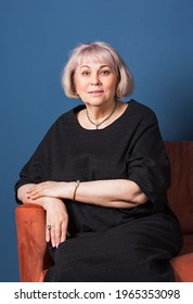 Confident Stylish European Mature Aged Woman Sitting At Chair, She Wearning At Black Dress. Portrait Of A Stylish Older Senior Business Woman, 60s Gray-haired Lady. Deep Blue Background.