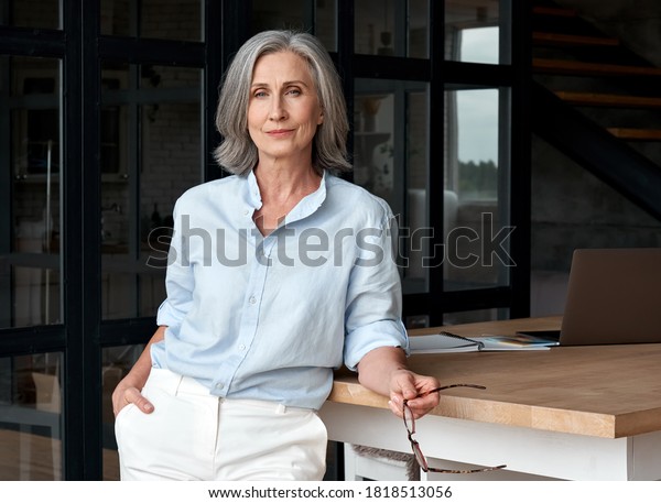 Confident stylish european mature middle aged\
woman standing at workplace. Stylish older senior businesswoman,\
60s gray-haired lady executive leader manager looking at camera in\
office, portrait.