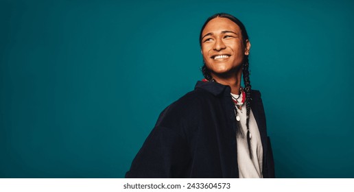 Confident, stylish young man with braided hair stands on a blue background. He wears casual clothing and a necklace, smiling happily while looking away. His ethnic heritage is Native American. - Powered by Shutterstock