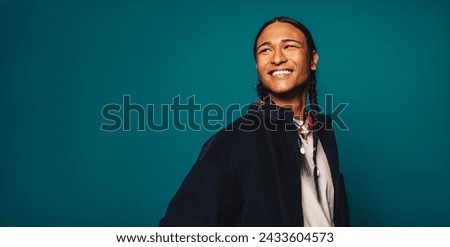 Confident, stylish young man with braided hair stands on a blue background. He wears casual clothing and a necklace, smiling happily while looking away. His ethnic heritage is Native American.