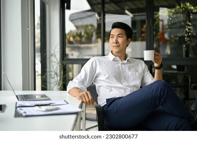 Confident and successful millennial Asian businessman or male CEO sits at his desk in modern office with a coffee cup in his hand. Businesspeople concept - Powered by Shutterstock