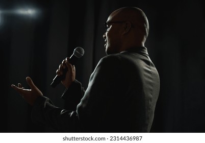 Confident successful speaker man talking on stage with spotlight strike through the darkness at corporate business event. Public speaker giving talk at conference hall. Stand up comedian. - Powered by Shutterstock