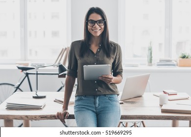 Confident and successful. Confident young woman in smart casual wear holding digital tablet and smiling while standing near her working place