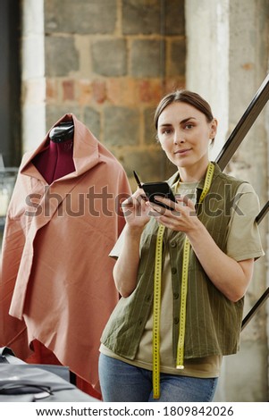 Confident tailor and owner of fashion studio looking at you while taking new orders from clients by table with sketches and unfinished clothes
