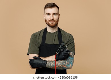 Confident tattooer master artist tattooed man wear green t-shirt apron hold hands crossed folded, machine black ink in jar, equipment for making tattoo art on body isolated on plain beige background - Shutterstock ID 2306086159