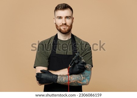 Confident tattooer master artist tattooed man wear green t-shirt apron hold hands crossed folded, machine black ink in jar, equipment for making tattoo art on body isolated on plain beige background