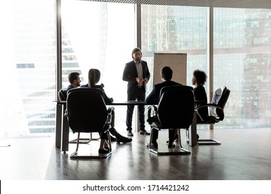 Confident Team Leader Giving Flip Chart Presentation At Corporate Meeting In Modern Boardroom With Panoramic Windows, Business People Sitting At Table, Listening To Coach Mentor Explaining Strategy