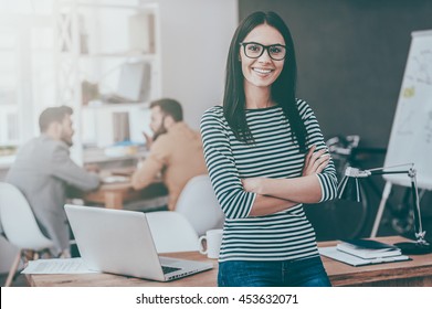 Confident Team Leader. Confident Young Woman Keeping Arms Crossed And Looking At Camera With Smile While Her Colleagues Working In The Background