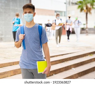 Confident Teen Boy In Medical Mask Walking Outside School Building On Autumn Day, Going To Lessons. Concept Of Necessary Precautions In COVID Pandemic