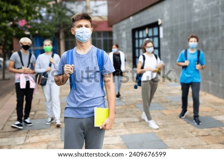 Confident teen boy in medical mask walking outside school building on autumn day, going to lessons. Concept of necessary precautions in COVID pandemic