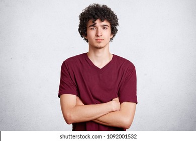 Confident teenager with crisp hair keeps hands crossed, looks directly into camera, listens attentively information, poses against white concrete wall. Self assured male student stands indoor