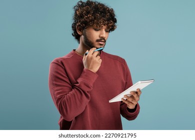 Confident thoughtful young Hindu man looking at the notepad, holding a pen at his chin while writing the list to do over isolated blue background. Portrait of a businessman, male entrepreneur, student - Powered by Shutterstock