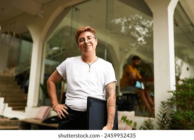 Confident transgender pro holds laptop, stands in modern workspace. Inclusive work culture, gender fluidity in tech. Office setting, casual style, diverse team, LGBTQ identity, coworking space. - Powered by Shutterstock