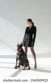 Confident Woman In Black Blazer Dress Looking Away Near Doberman Dog On Grey Background With Shadows