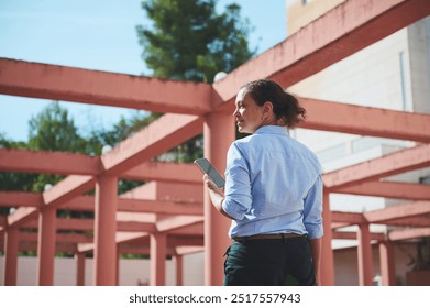 A confident woman in a blue shirt holds a smartphone while walking under pink beams in an outdoor setting, capturing a moment of urban exploration and modern connectivity. - Powered by Shutterstock