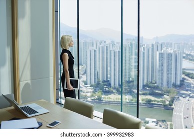 Confident woman business owner with digital tablet in hand is thinking about future meeting with investors, while is watching on developed New York city view with tall skyscrapers outside the window