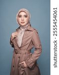 A confident woman, draped in a sophisticated taupe trench coat, showcasing a beautiful patterned hijab. A contemporary portrayal of modest and fashionable attire.