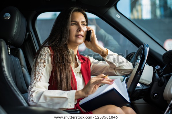 Confident woman executive director in formal wear\
talking via smartphone and using diary while sitting in automobile\
before work day in company. Female financier having cell telephone\
conversation 