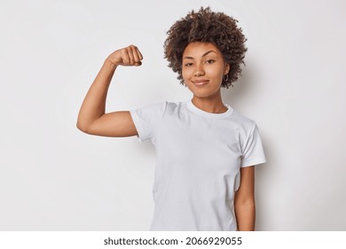 Confident woman feels strength and power raises arm flexes biceps and looks proud of her own achievements has strong muscles wears casual t shirt isolated over white background brags with fit body