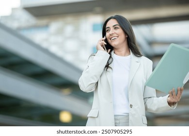 A confident woman in a light suit is engaged in a phone conversation, smiling while holding folders outside a modern building. - Powered by Shutterstock